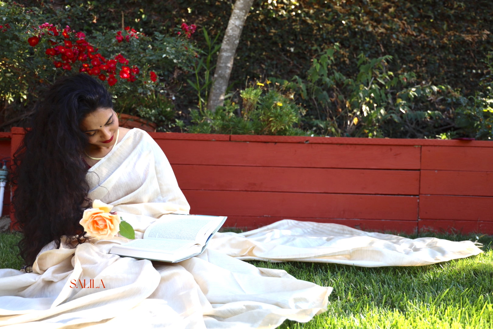 Salila reading the Ashtanga Hridayam in her backyard while wearing a while saree, and feeling cozy. Her long hair flows with the breeze.